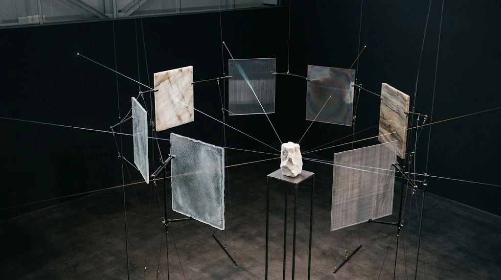Vertical panels of different materials and thin connections hang around a small stone object in the centre.